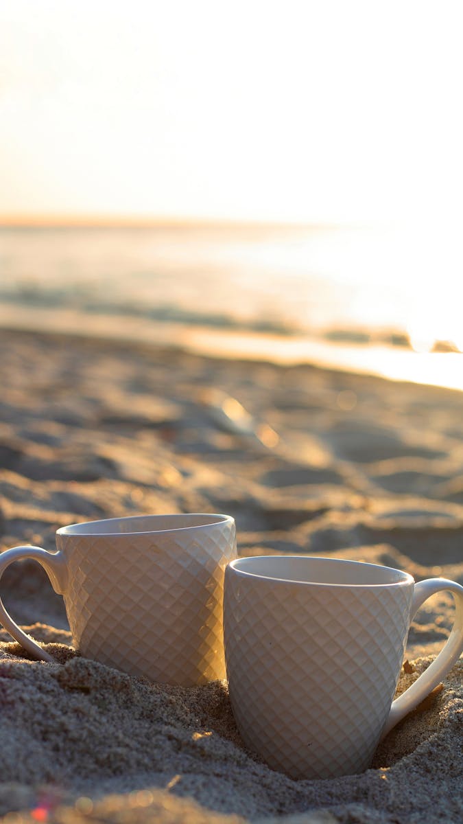 Two mugs on a sandy beach, warmly lit by the sunrise, creating a cozy, tranquil ambiance.