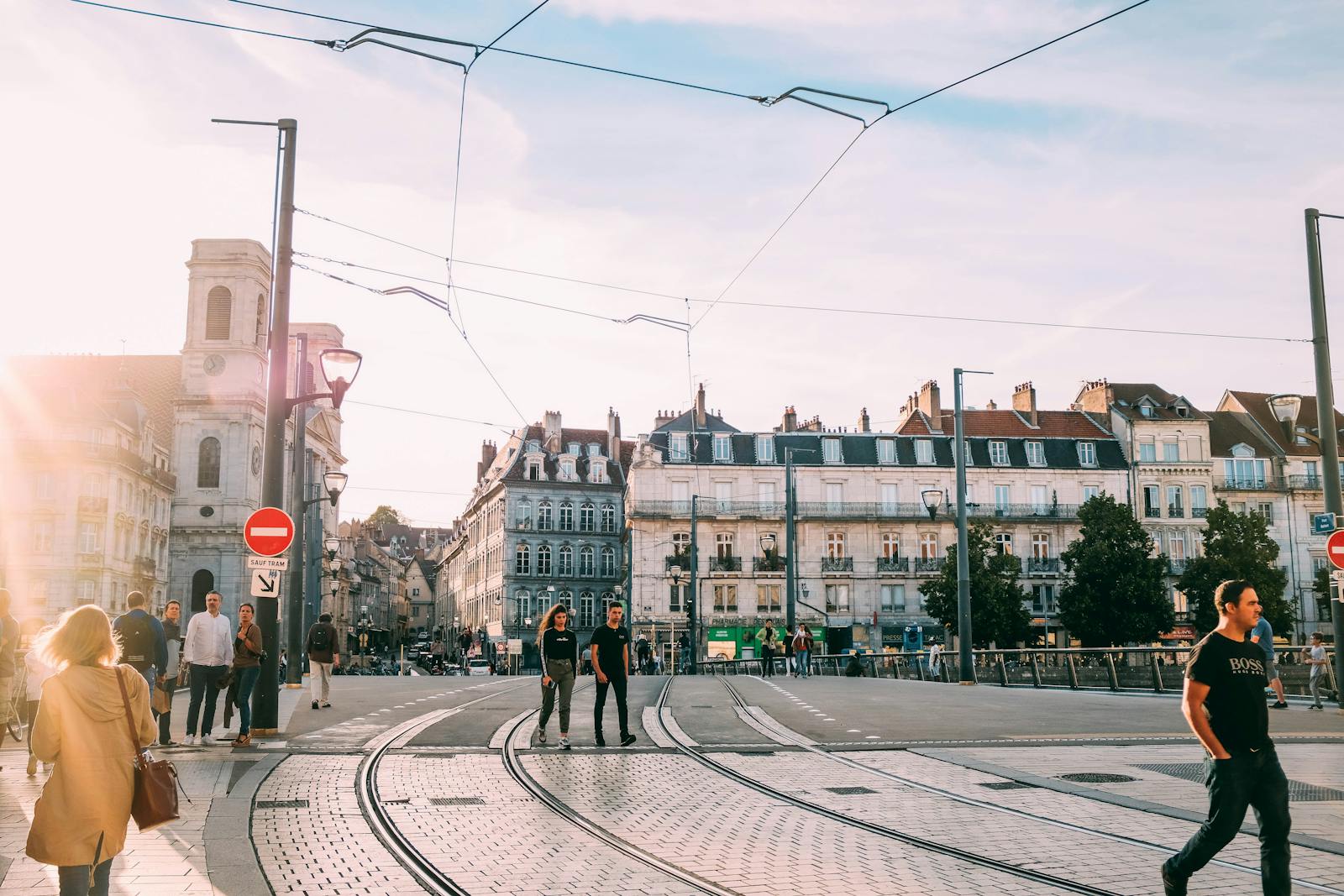 People walking across a sunny tramway intersection in a historic European city.