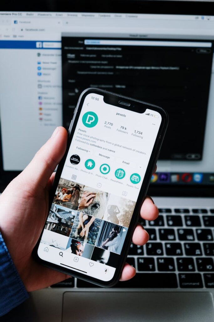 Hand holding smartphone displaying social media app with laptop in the background.