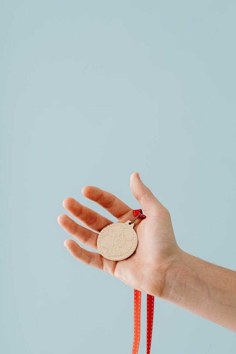 A close-up shot of a hand holding a gold medal with a red ribbon against a light blue background.