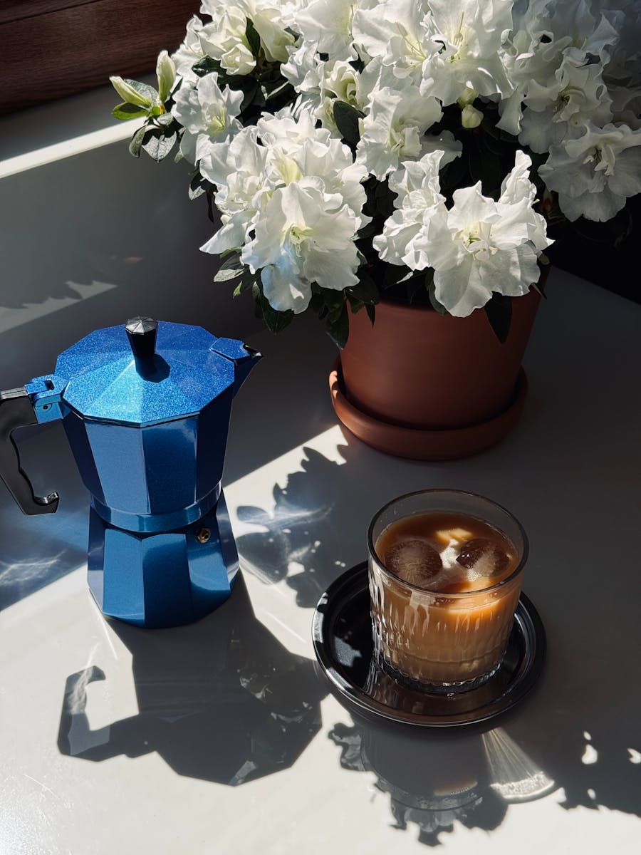 Elegant blue moka pot beside iced coffee and blooming azaleas in sunlight.
