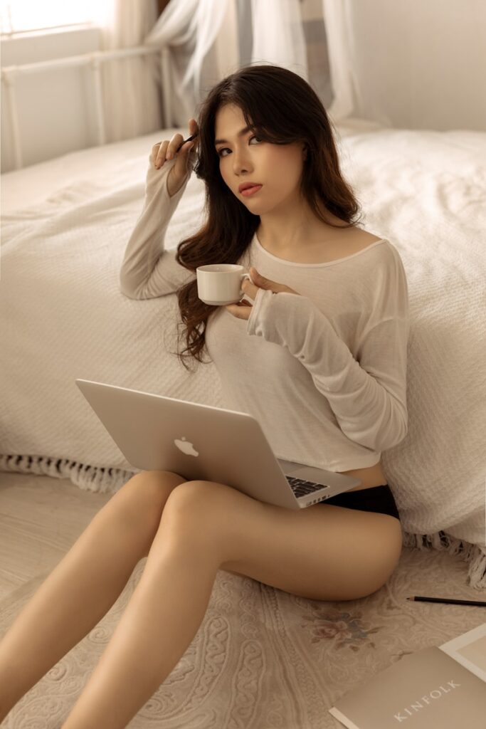 A young woman in white sweater sitting on bed with her laptop
